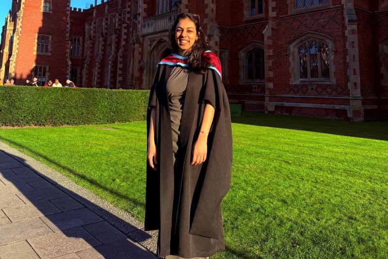 smiling woman with long wavy black hair, wearing a black graduation gown, standing outdoors with lawn and a large red brick three storey building behind her